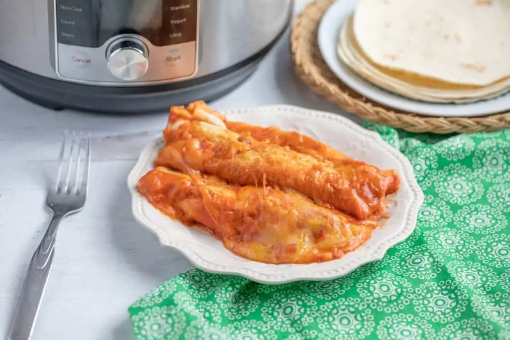 Instant Pot cheesy beef enchiladas on a white plate with the Instant Pot and tortillas in the background, decorative green kitchen cloth and silver fork on the countertop.