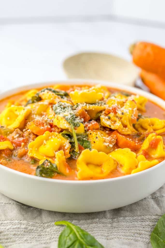 Close up of a bowl of Slow Cooker Tortellini Soup on a grey kitchen cloth, carrots and wooden spoon in the background.