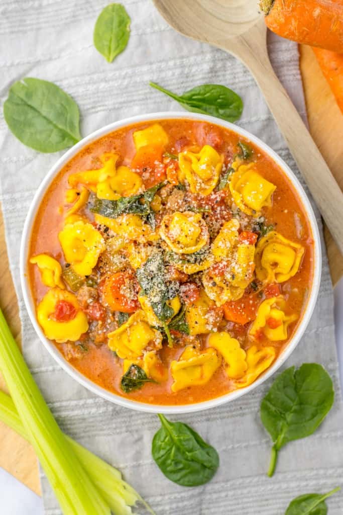 Overhead view of a bowl of Slow Cooker Tortellini Soup on a grey kitchen cloth with a wooden spoon, carrots, celery sticks, and baby spinach scattered around. 