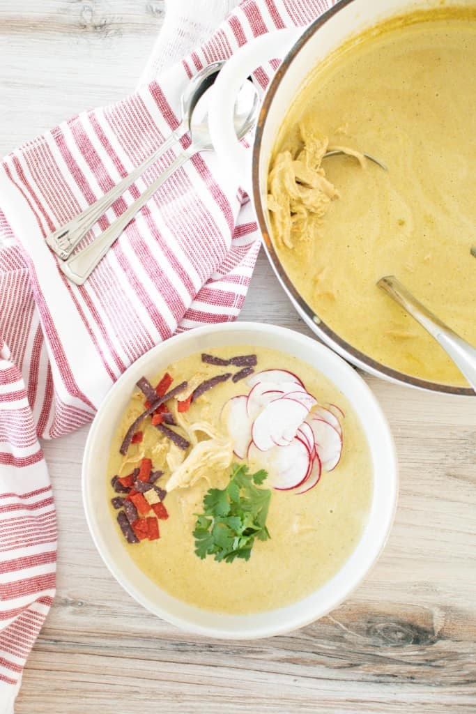 Overhead view of creamy chicken poblano soup in a white soup bowl, topped with garnish, on a wooden countertop with red-striped cloth and large pot full of soup in the background.