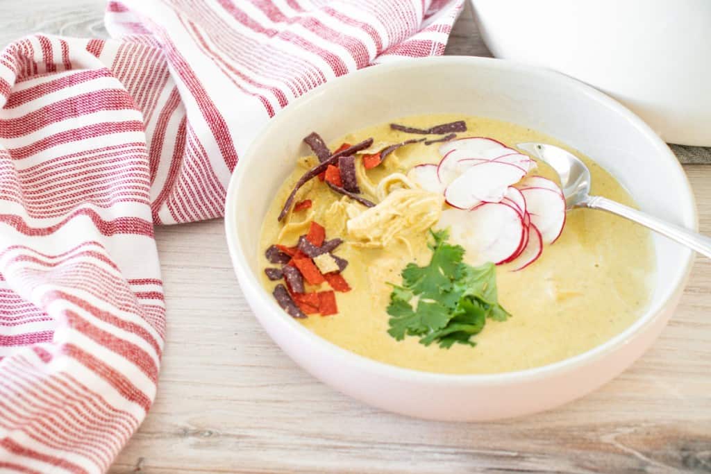 Creamy chicken poblano soup in a white soup bowl, topped with garnish, on a wooden countertop with red-striped cloth and large pot full of soup in the background.