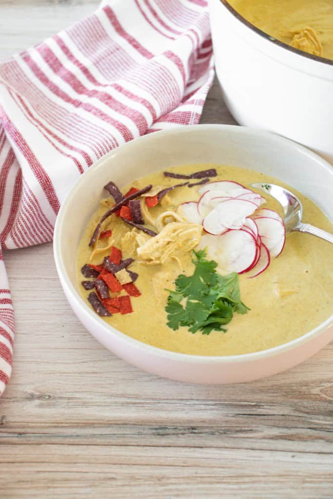 Creamy chicken poblano soup in a white soup bowl, topped with garnish, on a wooden countertop with red-striped cloth and large pot full of soup in the background.