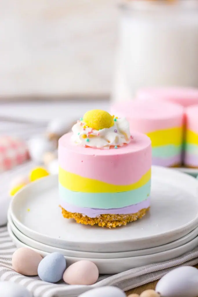 A no bake Easter mini cheesecake on a stack of white serving plates, a striped grey cloth and Easter egg candies displayed in front with more mini cheesecakes in the background.
