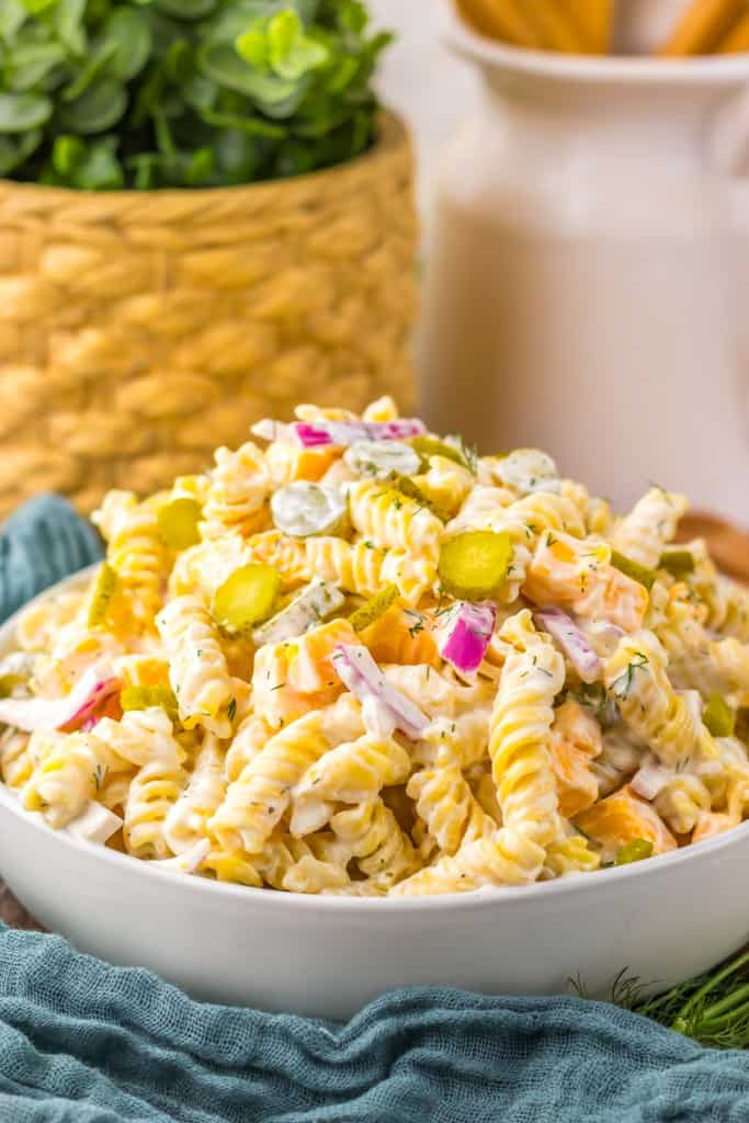 Pickle pasta salad in a white serving bowl, blue kitchen cloth and kitchen utensils with fresh dill on a wooden surface. 