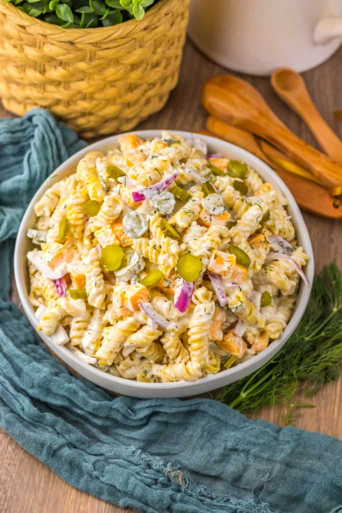 Overhead view of pickle pasta salad in a white serving bowl, blue kitchen cloth and kitchen utensils with fresh dill on a wooden surface. 
