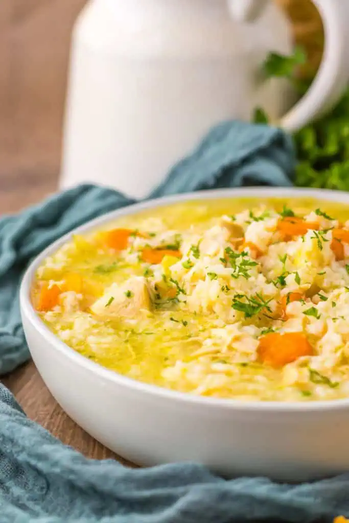 Slow cooker chicken and rice soup in a white serving bowl with a blue kitchen cloth, fresh parsley and cooking utensils in the background.
