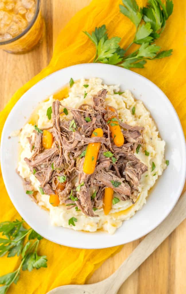 Overhead view of slow cooker garlic herb pot roast on a white dinner plate with a wooden serving spoon, fresh parsley, and a drink in the background.