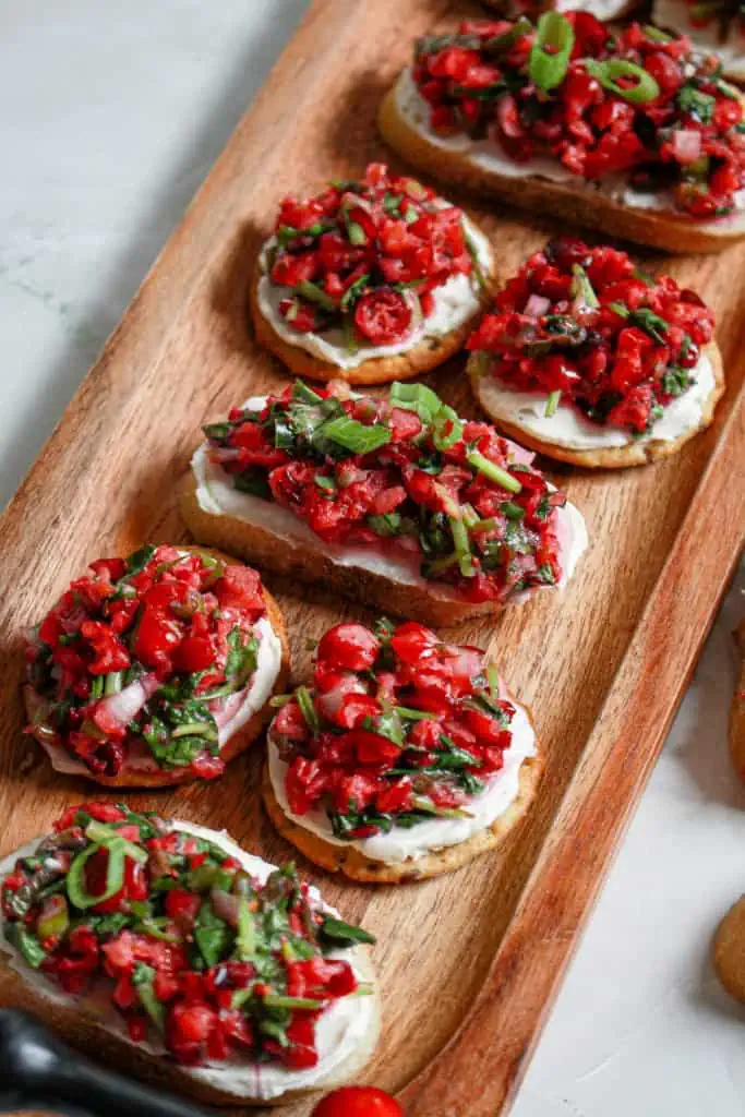 A wooden platter board loaded with cranberry jalapeno salsa appetizers, using crackers and bruschetta as the bases with a spread of cream cheese.