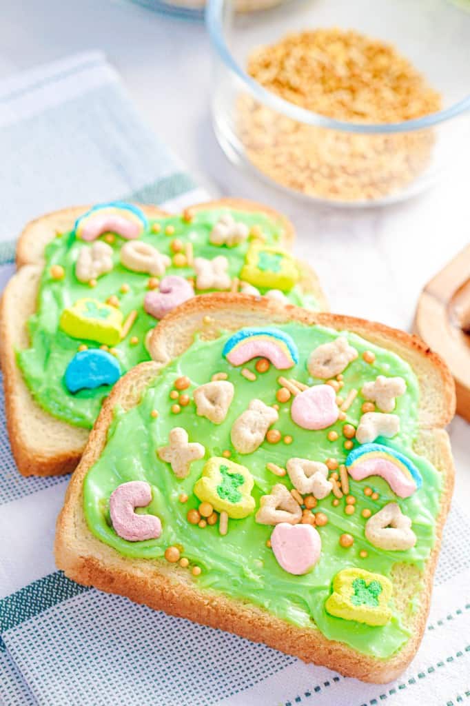 Two slices of leprechaun toast with Lucky Charms cereal and gold sprinkles on a marble countertop and decorative kitchen cloth. 
