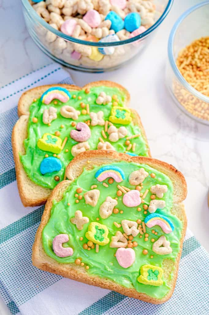 Two slices of leprechaun toast with Lucky Charms cereal and gold sprinkles on a marble countertop and decorative kitchen cloth. 