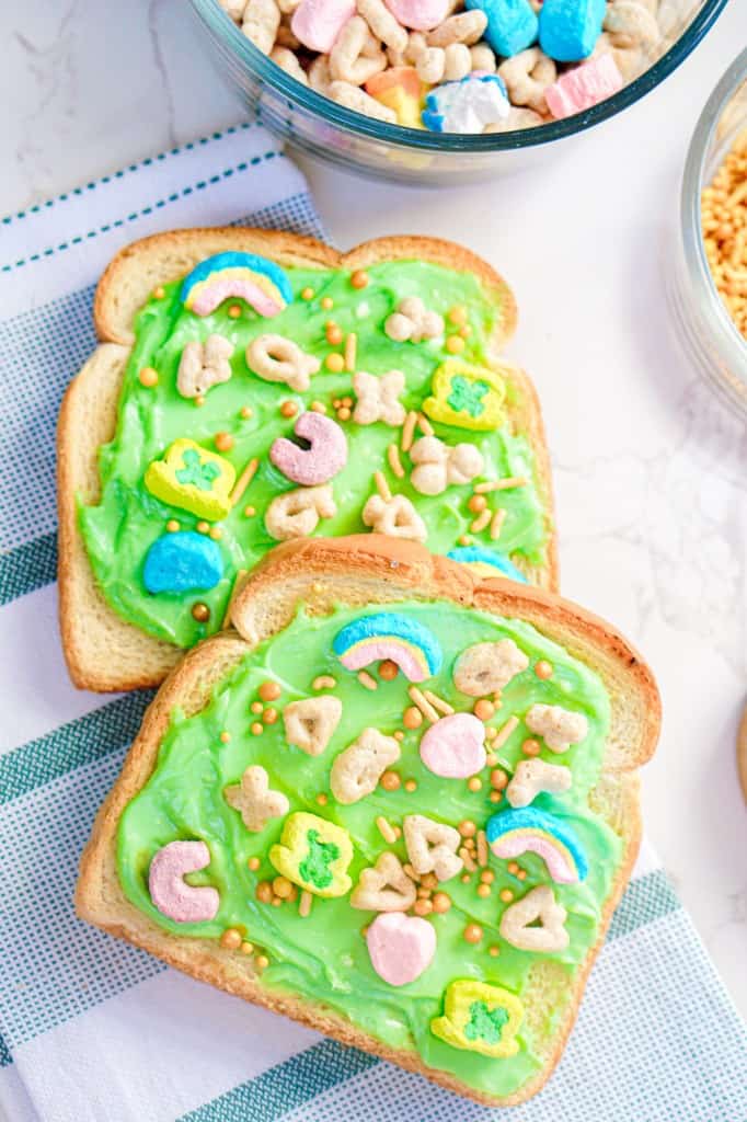 Close up of two slices of leprechaun toast with Lucky Charms cereal and gold sprinkles on a marble countertop and decorative kitchen cloth. 