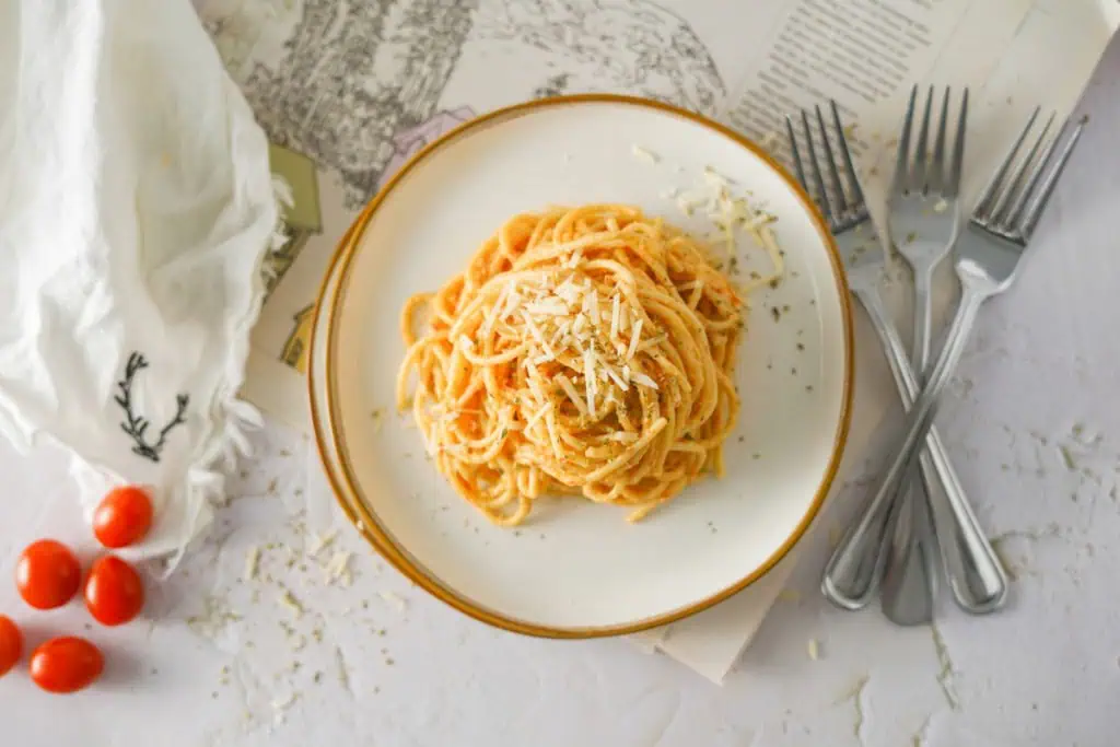 Overhead view of creamy burrata pasta on a serving plate, topped with grated Parmesan cheese. Silver forks, loose cherry tomatoes, and a kitchen cloth surrounding the dish.