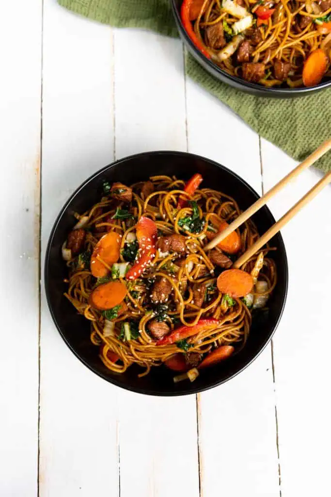 Overhead view of Instant Pot Pork Lo Mein in a black bowl with two chop sticks. Additional serving in bowl with green kitchen cloth in the upper corner of the image.