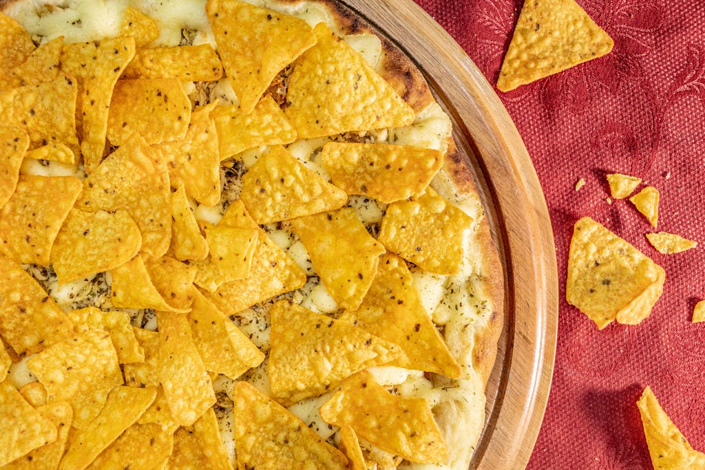 A top-down view of a cheesy pizza generously topped with crunchy nachos on a wooden platter.