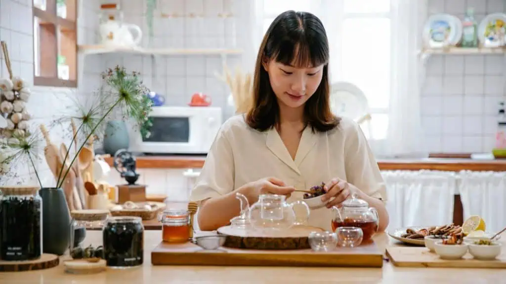 Woman preparing herbal tea with herbs and spices in a cozy kitchen setting filled with natural light.