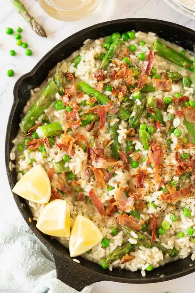 Over head view of creamy asparagus risotto in a cast iron skillet with crumbled bacon and topped with grated parmesan and lemon wedges
