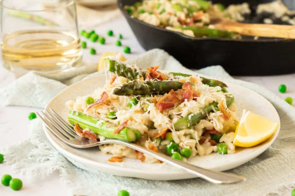 creamy asparagus risotto on a cream colored plate with crumbled bacon and topped with grated parmesan and slice of lemon