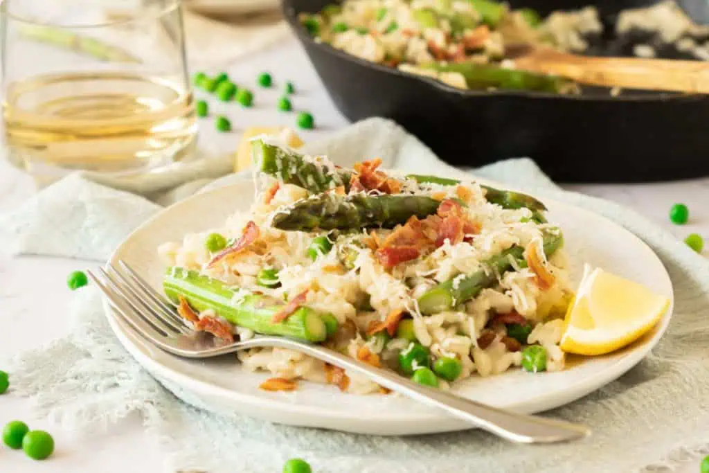 creamy asparagus risotto on a cream colored plate with crumbled bacon and topped with grated parmesan and slice of lemon