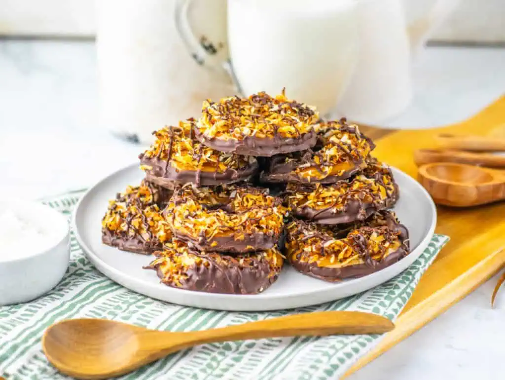 side view of homemade Samoas cookies on a white plate showing the toasted coconut layer, dark chocolate-dipped base, and thin chocolate drizzle over the top.