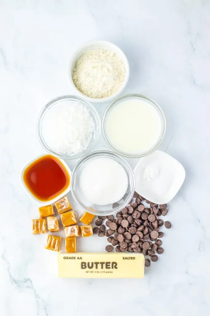Homemade Samoas cookie ingredients arranged overhead on a white surface: butter, sugar, flour, milk, vanilla, sweetened shredded coconut, wrapped caramels, and a dark chocolate bar.