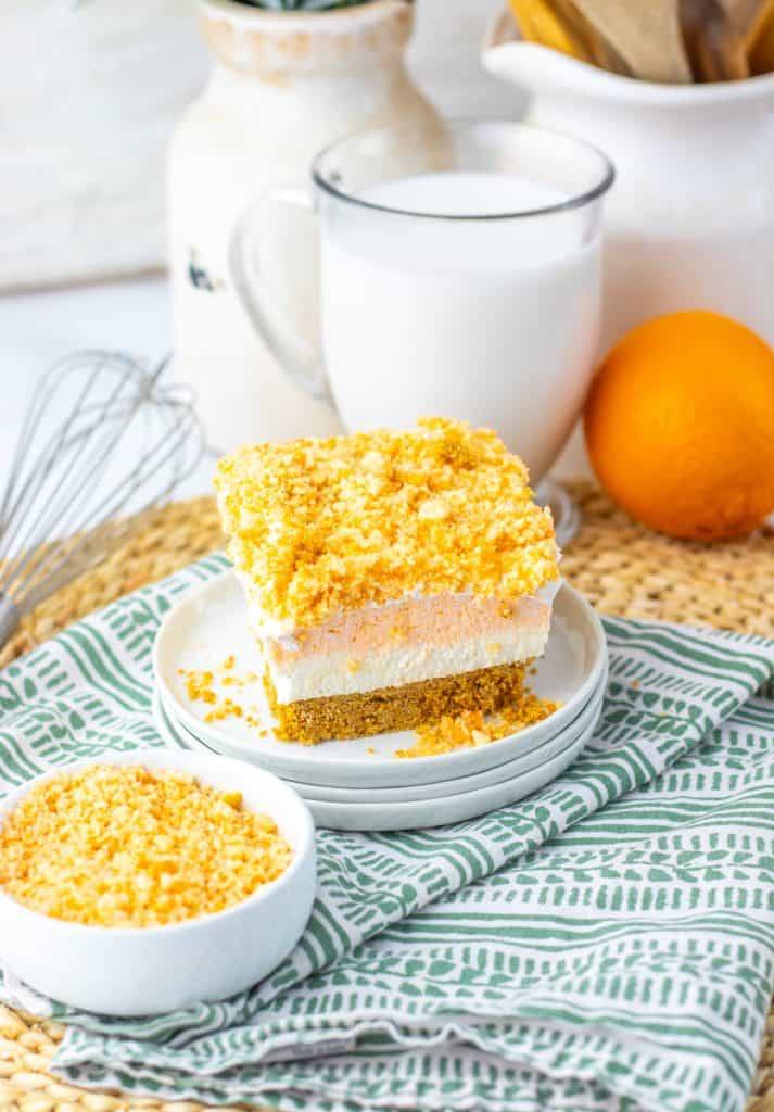 Orange vanilla Oreo crunch topping being sprinkled over the white whipped topping layer of the bars in an 8x8 parchment-lined pan, with a bowl of the orange crumb mixture alongside.
