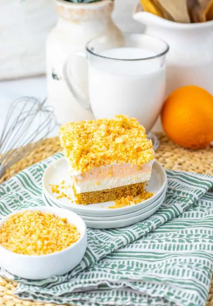 Orange vanilla Oreo crunch topping being sprinkled over the white whipped topping layer of the bars in an 8x8 parchment-lined pan, with a bowl of the orange crumb mixture alongside.