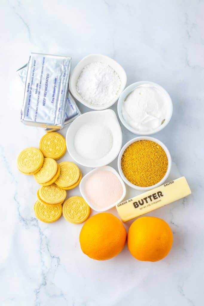 Orange creamsicle crunch bar ingredients arranged overhead: graham crackers, butter, cream cheese blocks, powdered sugar, orange, orange jello boxes, Cool Whip, and vanilla Oreos on a white surface.
