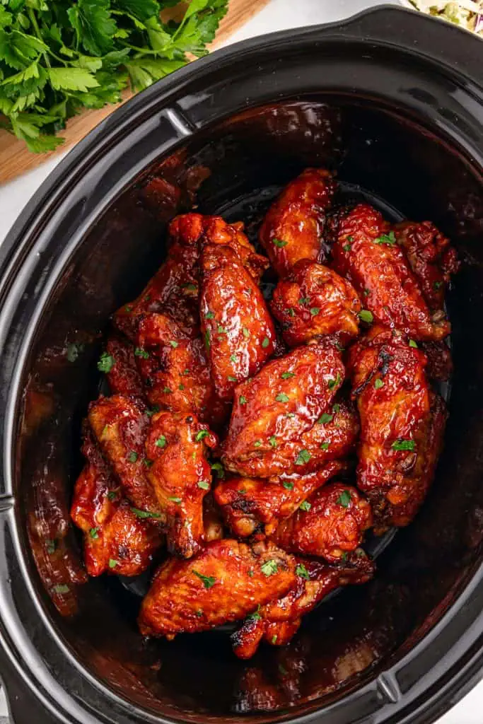 Close-up of tender BBQ chicken wing coated in thick smoky sauce with crispy caramelized edges in a black slow cooker.
