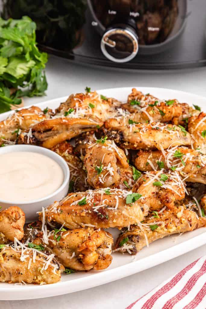 Chicken wings, grated parmesan, minced garlic, parsley, and seasonings arranged on a white plate in front of a black slow cooker