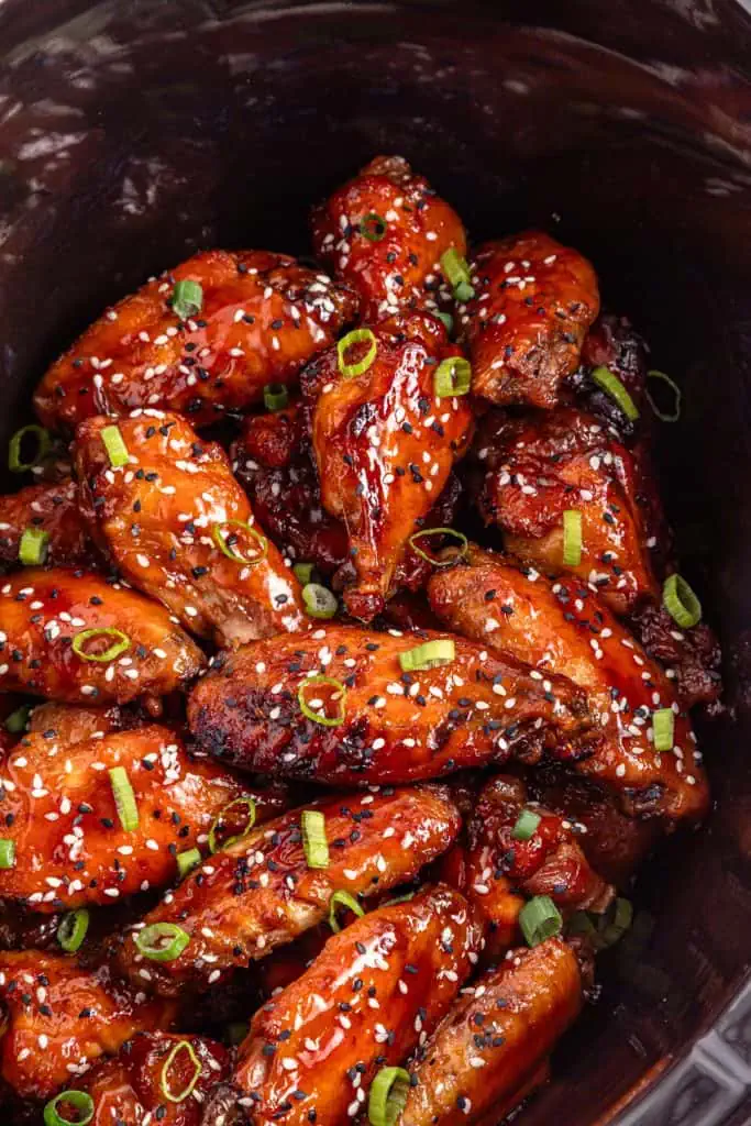 Close-up of teriyaki chicken wing showings a thick, glossy, caramelized teriyaki glaze with sesame seeds and sliced green onions on top in a black slow cooker.