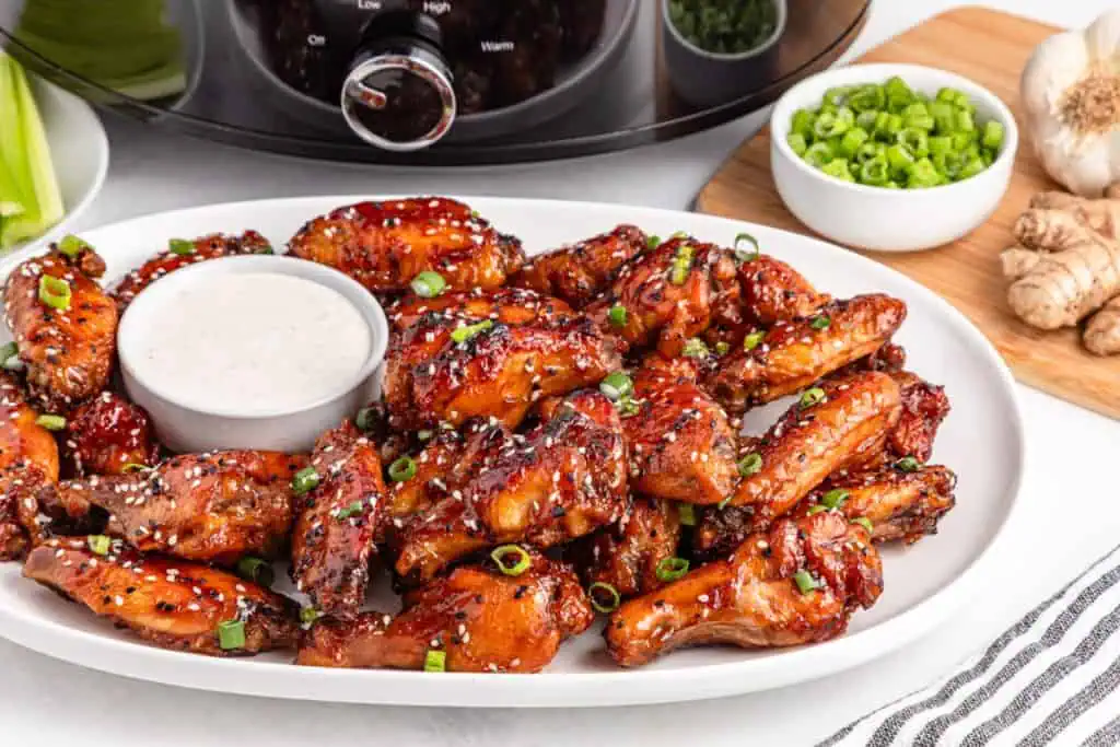 Sticky slow cooker teriyaki chicken wings piled on a white serving plate, garnished with sliced green onions and toasted sesame seeds with a small bowl of teriyaki dipping sauce alongside.
