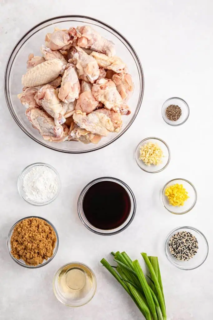 Slow cooker teriyaki chicken wings ingredients arranged overhead: chicken wings, soy sauce, brown sugar, rice vinegar, cornstarch, fresh garlic cloves, fresh ginger, black pepper, green onions, and sesame seeds.