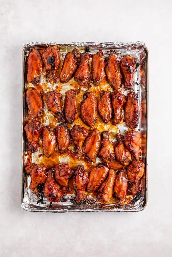 Teriyaki glaze being brushed onto slow-cooked chicken wings on a foil-lined baking sheet before going under the broiler, with a silicone brush and bowl of sauce in frame.