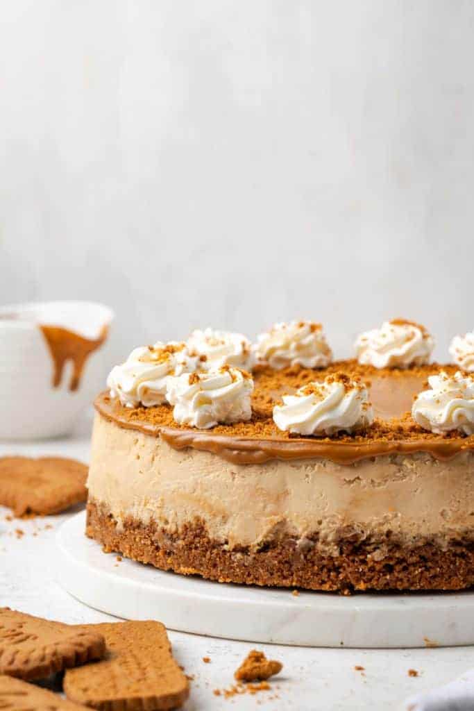 A whole Biscoff cheesecake on a white cake stand with a glossy Biscoff spread topping, scattered Biscoff cookie crumbs, and piped whipped cream rosettes around the edge.
