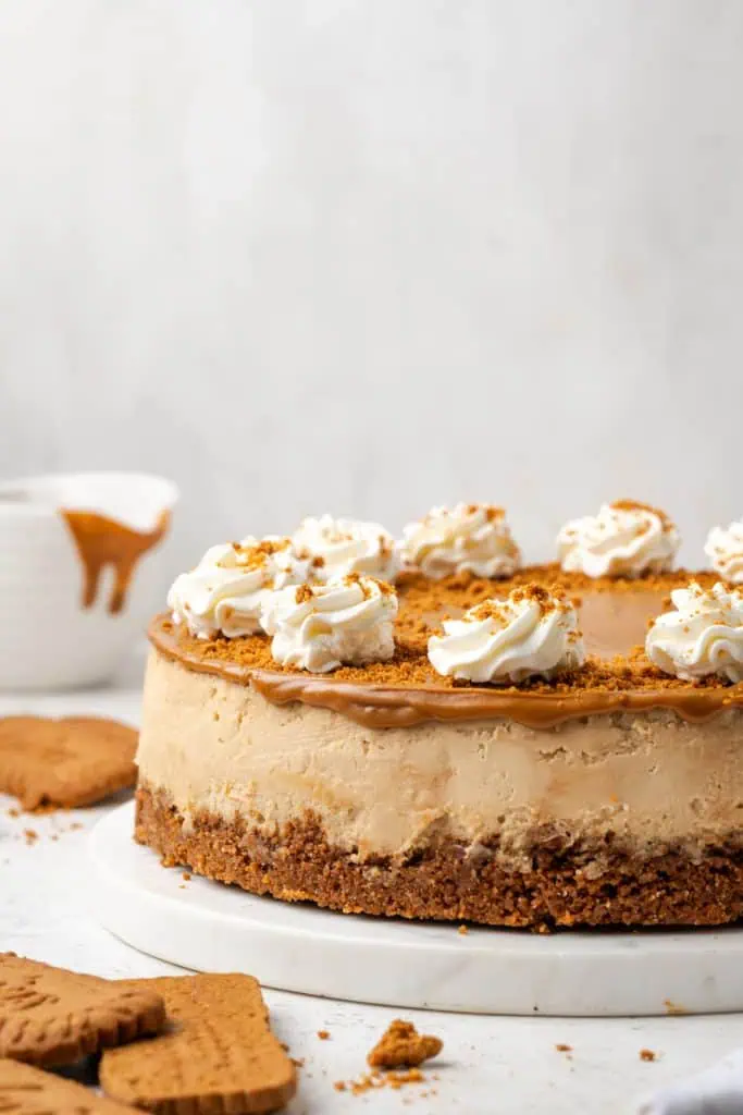 A whole Biscoff cheesecake on a white cake stand with a glossy Biscoff spread topping, scattered Biscoff cookie crumbs, and piped whipped cream rosettes around the edge.