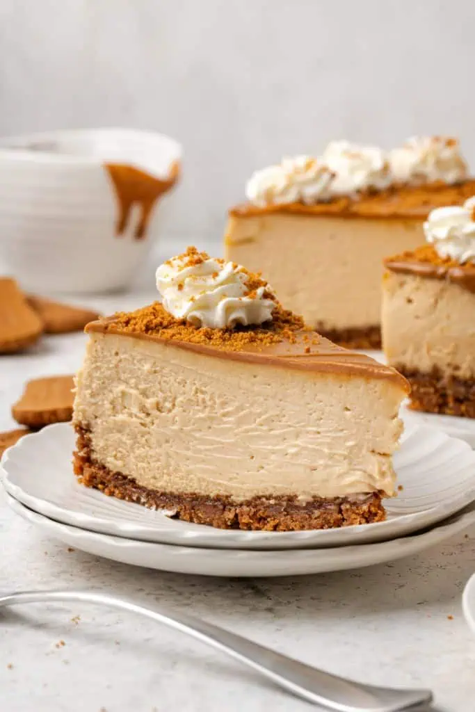 A single slice of Biscoff cheesecake on a white plate showing the dark Biscoff cookie crust, the creamy beige filling, and the glossy amber Biscoff glaze on top.