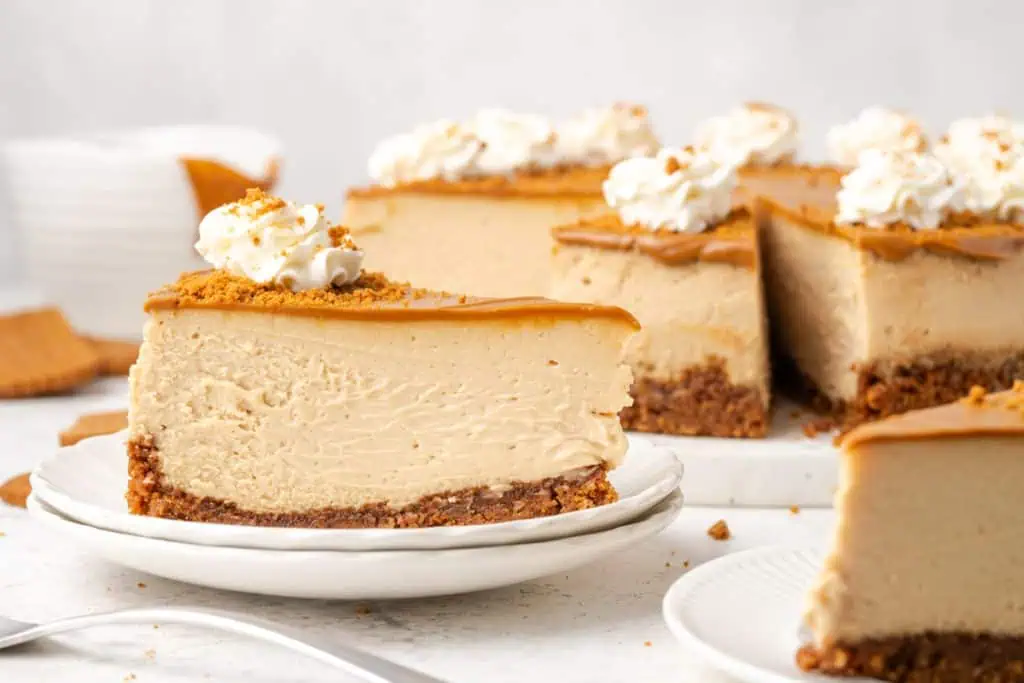 A slices of Biscoff cheesecake on a white plate showing the dark Biscoff cookie crust, the creamy beige filling, and the glossy amber Biscoff glaze on top.
