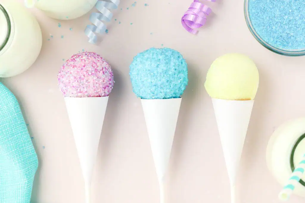 Colorful snow cone cake pops coated in rainbow sanding sugar and served in white paper cones arranged on a light pink surface