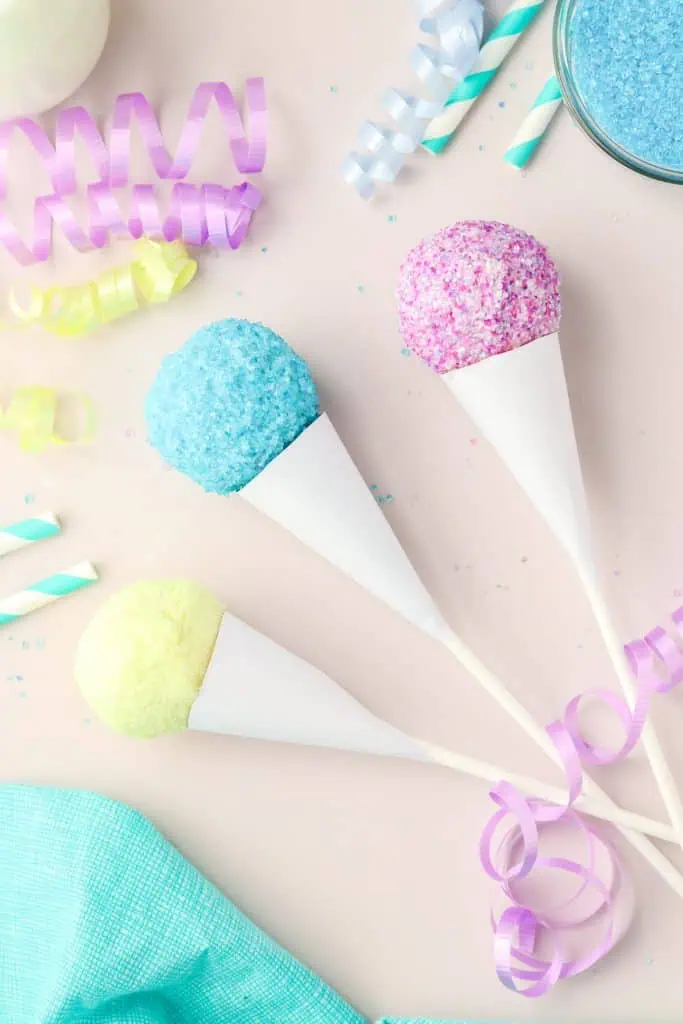 Overhead flatlay of snow cone cake pop ingredients including a baked 9x13 cake, white candy melts, colorful sanding sugar sprinkles, frosting, candy sticks, and white card stock arranged on a white surface