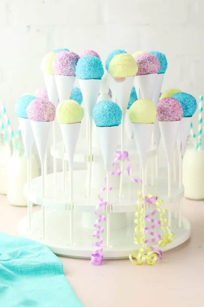 Three snow cone cake pops in handmade white paper cones standing upright, decorated with colorful sanding sugar, styled on a bright summer party table