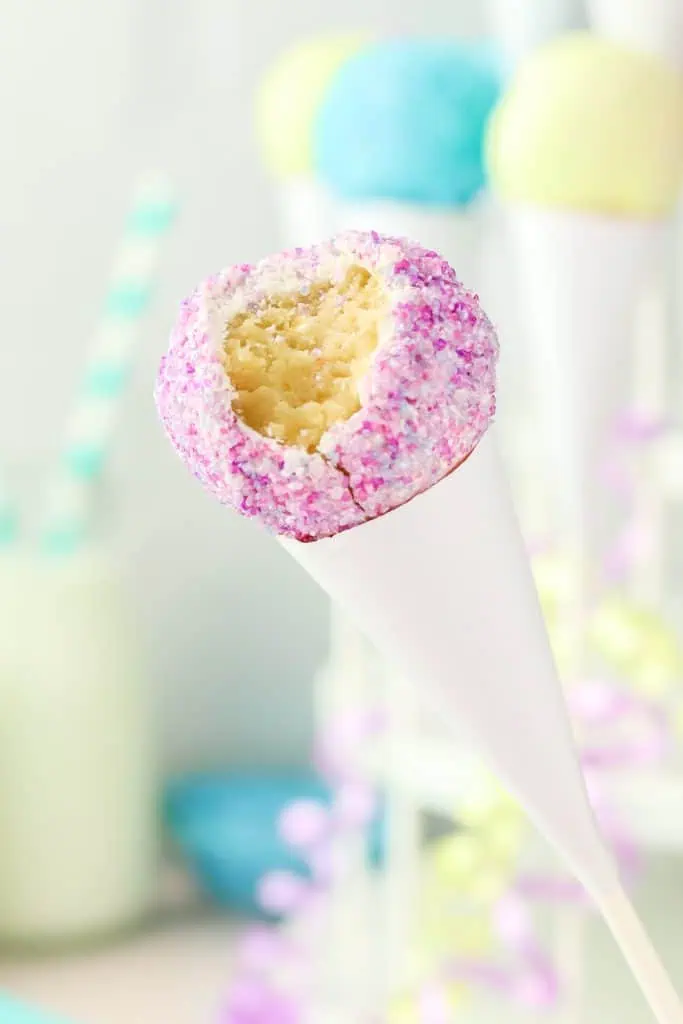 Close-up of a snow cone cake pop covered in bright pink and blue sanding sugar crystals on a candy stick with a bite taken out of it