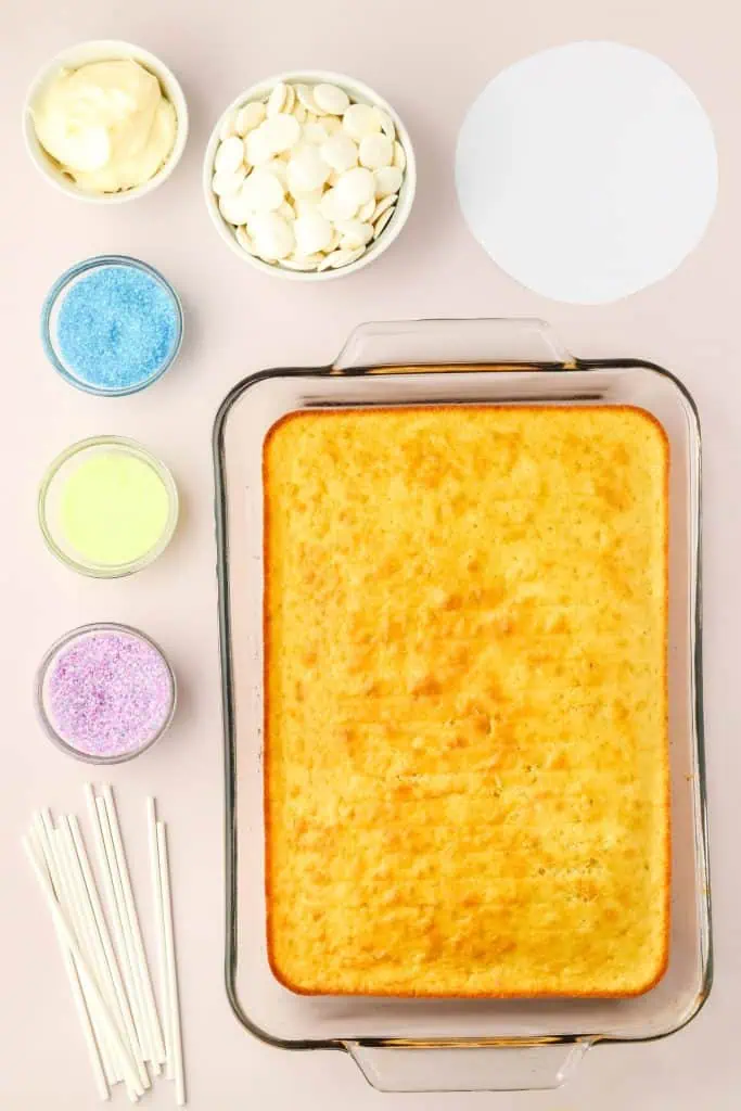 All of the ingredients you need to make these super fun summer dessert treats of snow cone cake pops with cake cooked in a glass dish, and sanding sugars in different bowls, and a bowl of white chocolate melts.