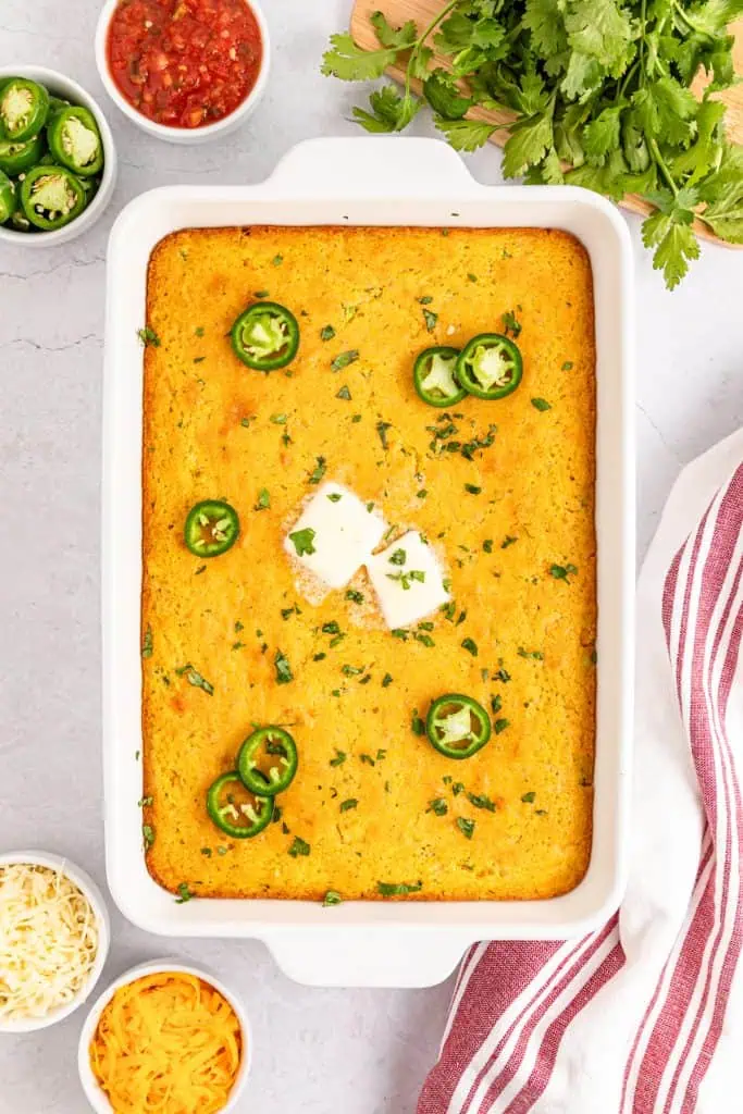an 9 x 13 pan of cheesy jalapeno cornbread with sliced jalapeños, sliced tabs of butter, and fresh parsley chopped on top to garnish on a white marble table