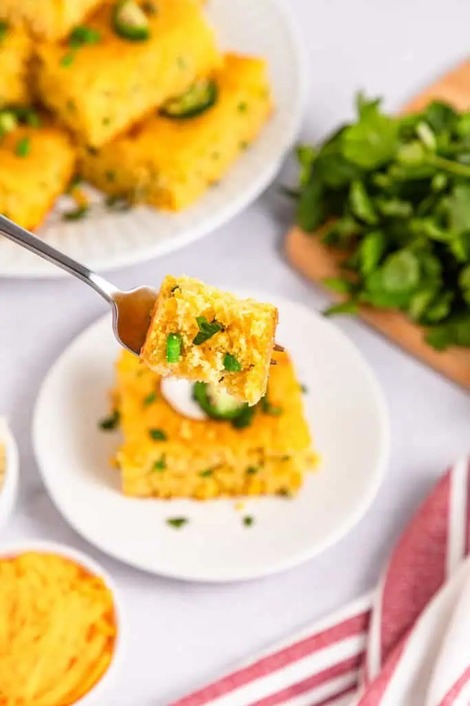 Golden jalapeno cornbread cut into squares in a 9x13 pan, topped with fresh cilantro and visible jalapeno slices on a white ceramic plate on a fork