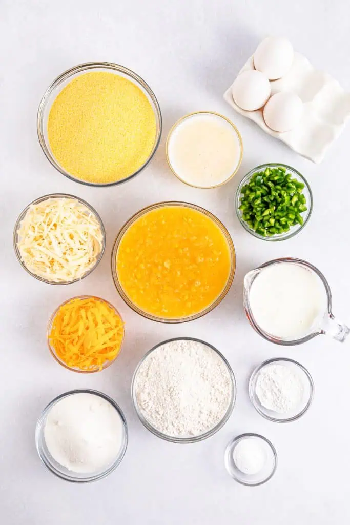Overhead flatlay of jalapeno cornbread ingredients including cornmeal, flour, buttermilk, eggs, cream corn, jalapenos, shredded cheese, and butter on a white surface