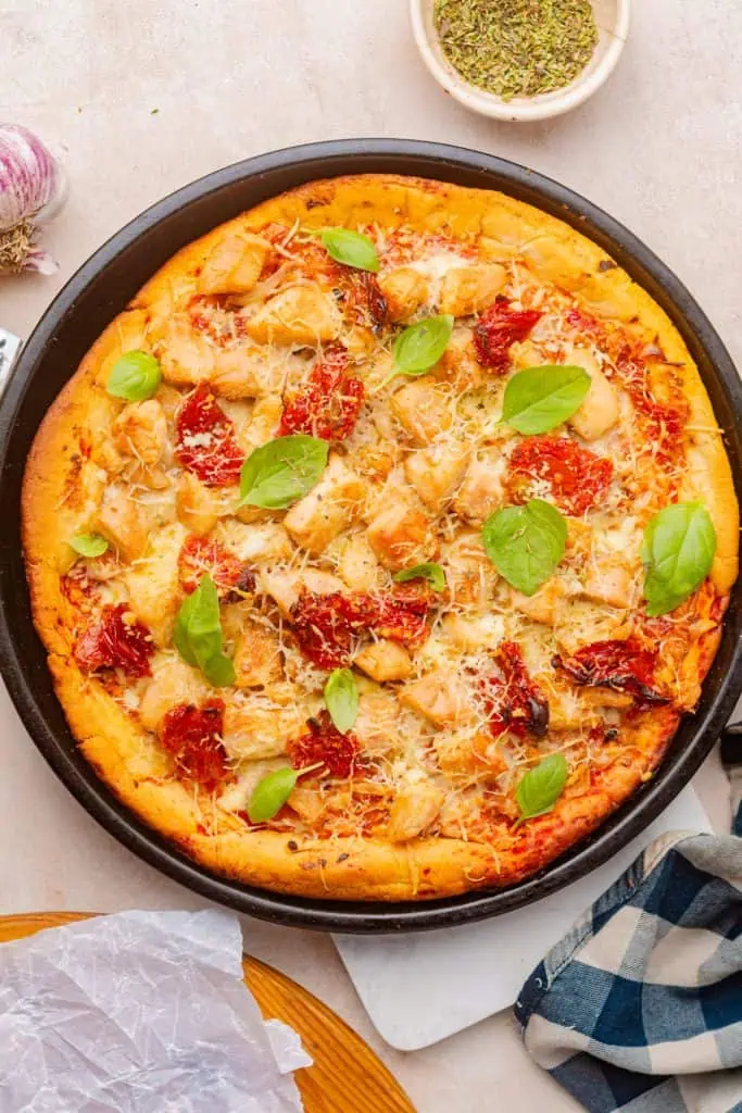 Marry me chicken pizza whole on a perforated pizza pan, cheese golden and bubbling, sun-dried tomatoes and chicken visible, fresh basil scattered over after baking