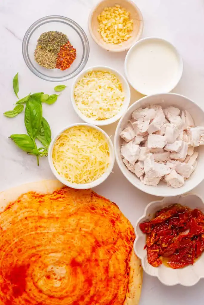 Par-baked pizza crust, chicken breast cubes, sun-dried tomatoes, heavy cream, mozzarella, Parmesan, garlic, fresh basil and dried herbs arranged on a wooden pizza board
