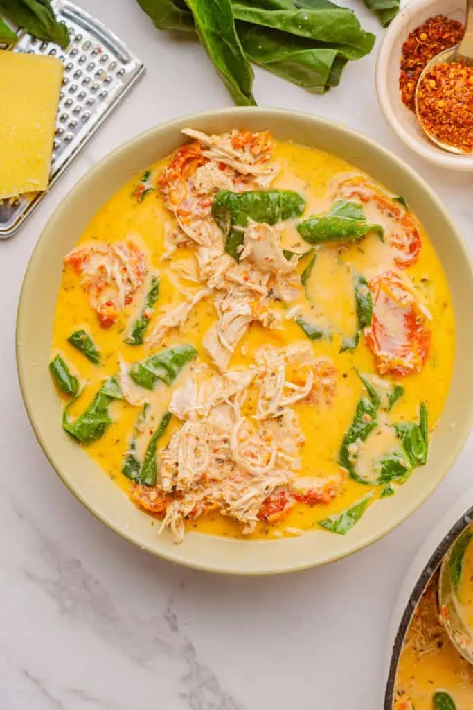 Close-up of marry me chicken soup surface showing golden cream swirl, vibrant green spinach leaves, and ruby-red sun-dried tomato pieces