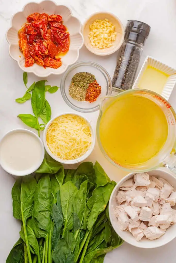 Shredded chicken, chicken stock, heavy cream, sun-dried tomatoes, baby spinach, garlic, Parmesan, and herbs arranged around a medium saucepan on a light wood surface