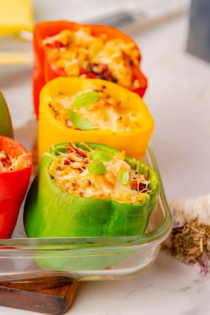 Three golden-topped marry me chicken stuffed peppers standing upright in a white baking dish, Parmesan crust deeply golden, fresh basil scattered over